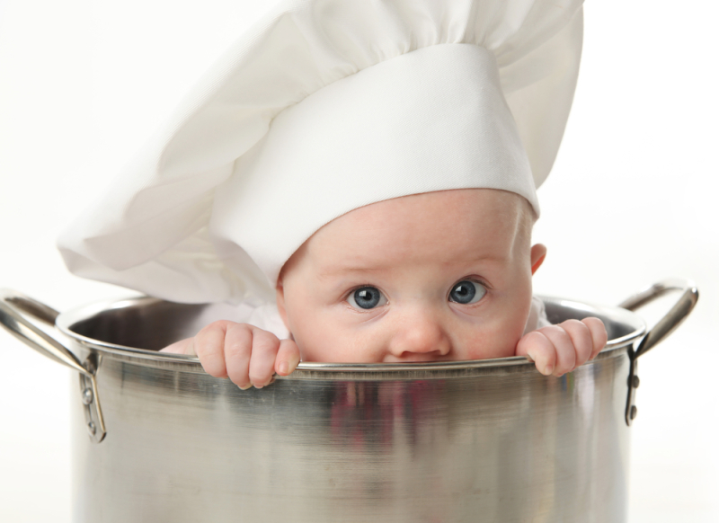 Close up portrait of a baby sitting wearing a chef hat sitting inside a large cooking stock pot, isolated on white
