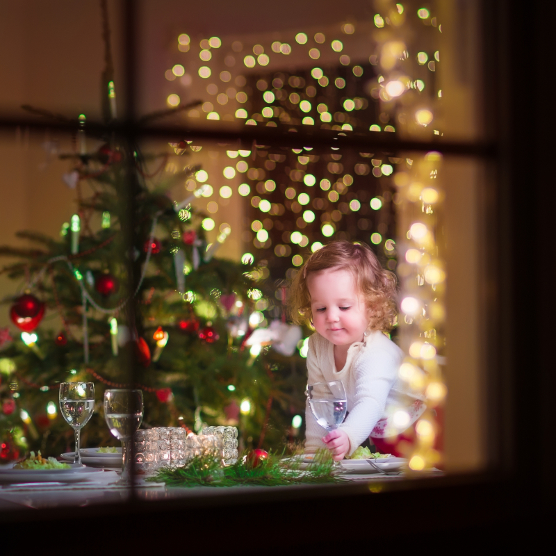 Cute curly toddler girl standing at a Christmas dinner table settling the dishes preparing to celebrate Xmas Eve, view through a window from outside into a decorated dining room with tree and lights 