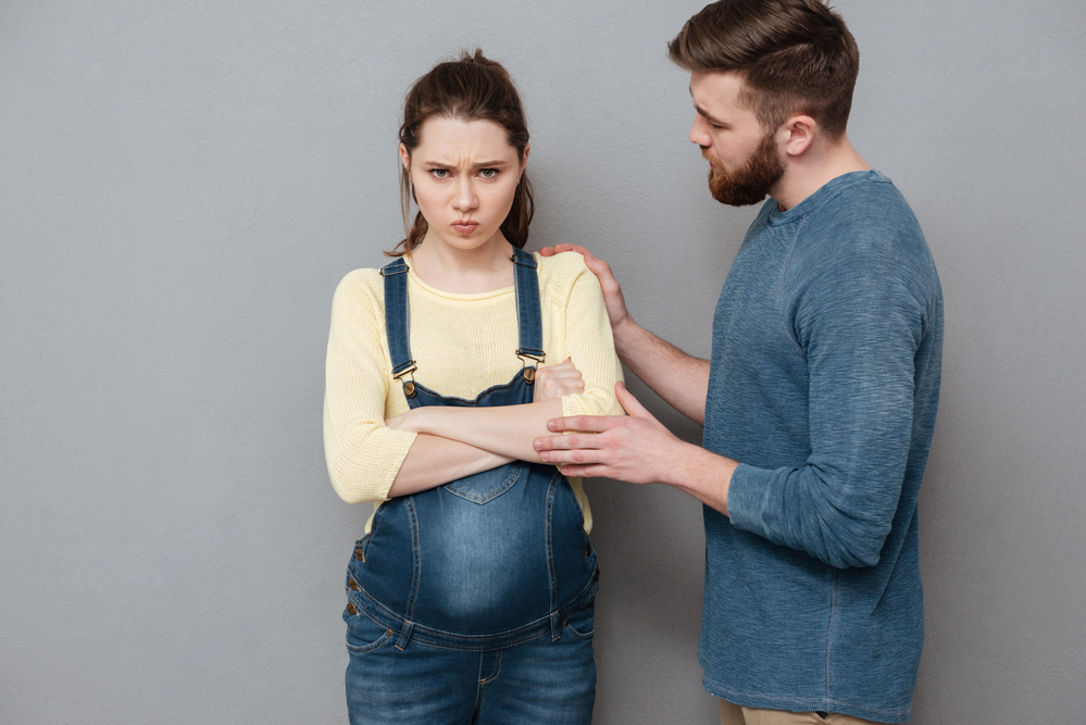 Image of pregnant angry woman standing near confused man isolated over grey wall.