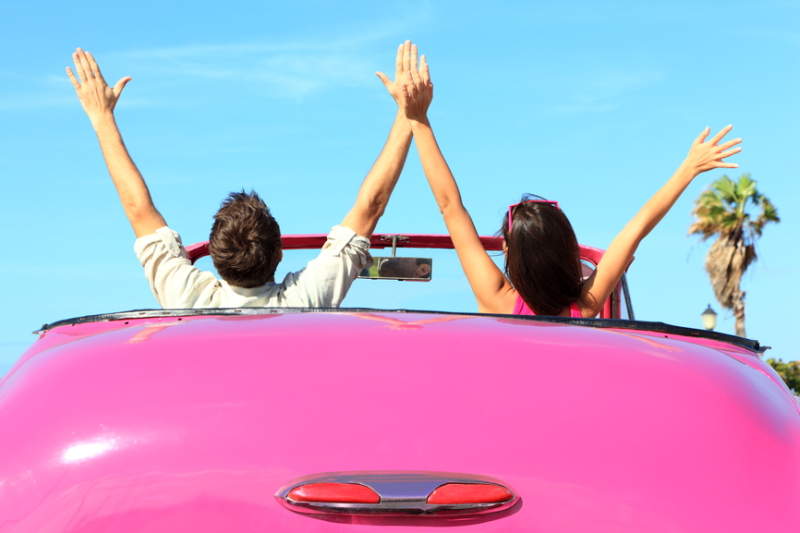 Freedom - happy free couple in car driving in pink vintage retro car cheering joyful wih arms raised. Friends going on road trip travel on summer day under sun blue sky.