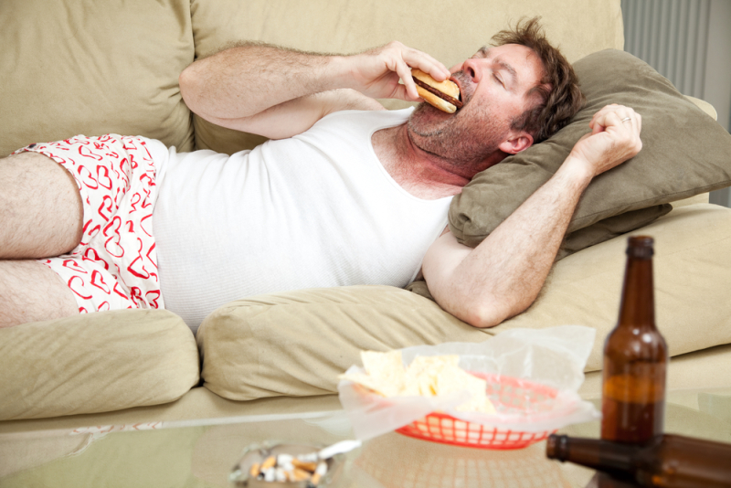 Unemployed middle aged man at home on the couch in his underwear, eating a hamburger, with a marijuana joing in the ashtray and beer bottles lying around.